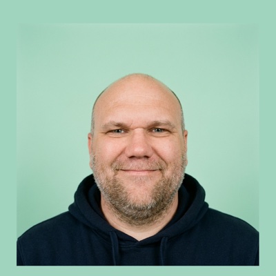 Alexander Ramm lächelt in die Kamera, dunkelblauer Kapuzenpullover vor grünem Hintergrund.