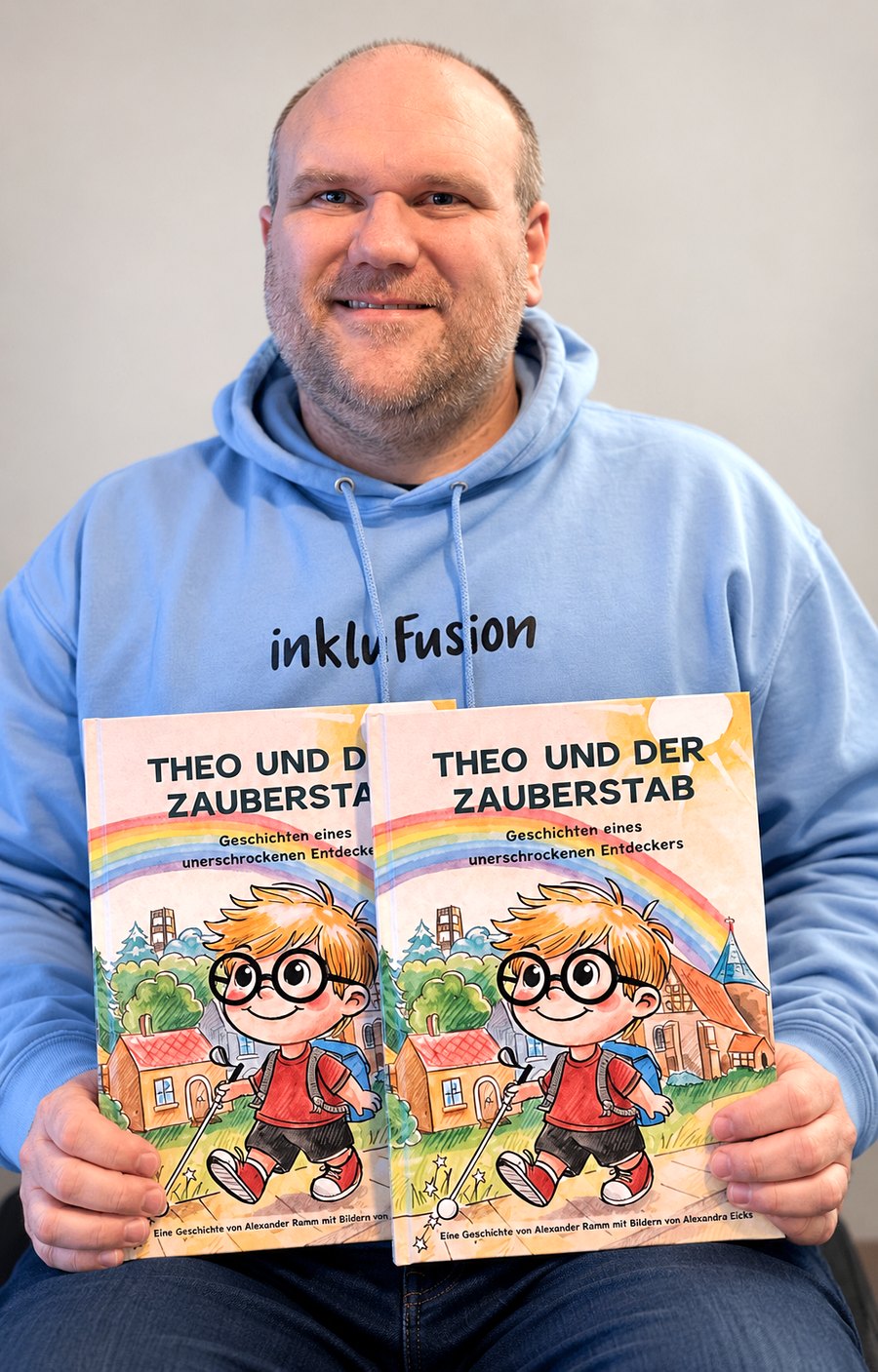 Alexander Ramm sitzt vor einer hellgrauen Wand. Er trägt einen hellblauen Kapuzenpullover mit dem Aufdruck „inkluFusion“ und hält zwei Exemplare seines Kinderbuchs „Theo und der Zauberstab“ vor sich. Er lächelt in die Kamera.